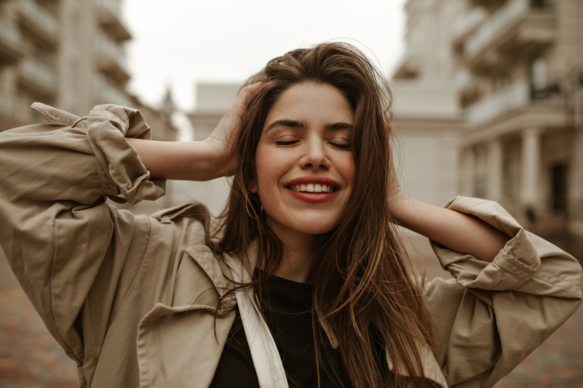 Emotional Happy Brunette Woman Smiles Sincerely and Ruffles Hair. Charming Woman in Beige Trench Co